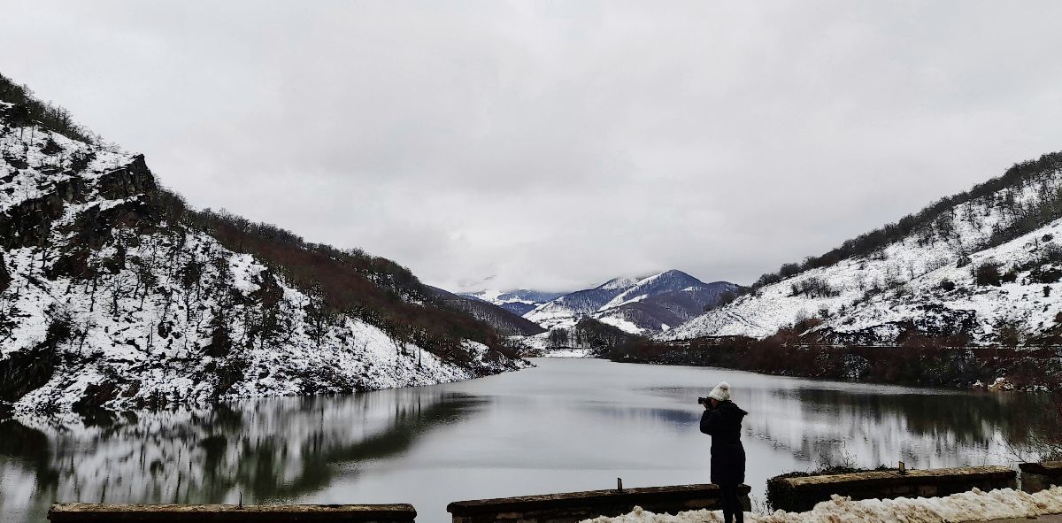 mujer haciendo foto con paisaje nevado y embalse con reflejo de montañas